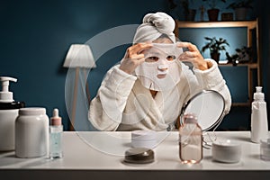 Beautiful lady applying cotton face mask sitting at table with various homemade cosmetics. Woman using cosmetic for