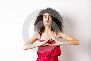 Beautiful lady with curly hair, red dress, showing heart symbol and pucker lips for kiss, love you gesture, standing on