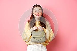 Beautiful lady holding her purse in hands near chest, smiling and looking excited at camera, waiting for something with