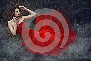 Beautiful lady in nrigh scarlet fishtail dress and vintage necklace sitting in the dry woods covered with smoke