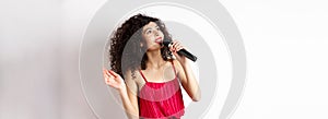 Beautiful lady in red dress singing songs in microphone, smiling and looking up, standing on white background