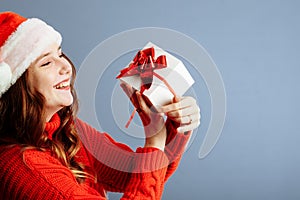 Beautiful lady in Santa hat is holding presents, looking at camera and smiling, isolated on gray background