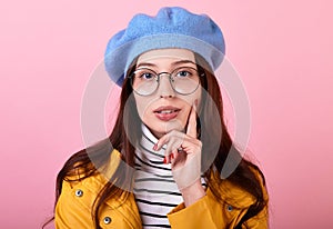 Beautiful lady in a stylish beret hat and a yellow raincoat with glasses shows dreams on a pink background