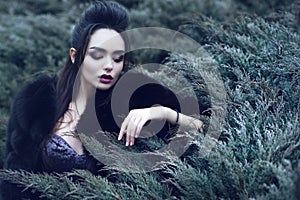 Beautiful lady wearing luxurious sequin dress and black sable fur coat sitting in coniferous bush and touching it with her fingers