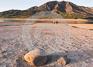 Beautiful landscape with setting sun: mountains, lake, cracked earth with stones -dried-up riverbend. Two people are standing in