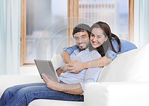 Beautiful latin couple in love lying together on living room sofa couch enjoying using digital tablet