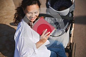 Beautiful Latin woman smiling looking aside, holding book and pushing baby pram with her sleeping child. Maternity leave