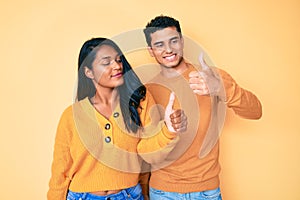 Beautiful latin young couple wearing casual clothes together looking proud, smiling doing thumbs up gesture to the side