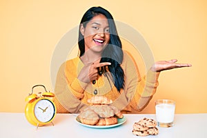 Beautiful latin young woman with long hair sitting on the table having breakfast amazed and smiling to the camera while presenting