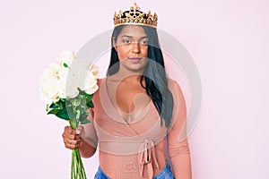 Beautiful latin young woman with long hair wearing princess crown and holding flowers thinking attitude and sober expression