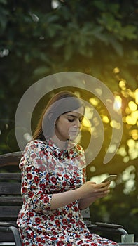Beautiful latin young woman in white floral design dress reading on her smartphone in the garden at sunset with the sun`s rays