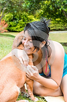 Beautiful latina girl sitting on the grass, very happy and with a big smile hugging her pet. mixed breed dog playing with her