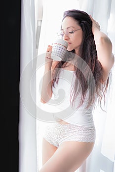 Beautiful Latina woman in white lingerie standing by a window and drinking coffee