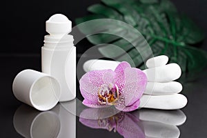 Beautiful lilac orchid flower and white roll-on deodorant with white stones and monstera leaves on black background