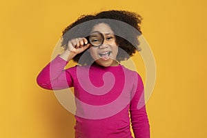 Beautiful little african girl looking through magnifying glass on yellow background