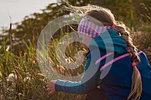 Beautiful little blond hair girl with long braid in blue hoody on wild blooming nature background.Child sitting alone.
