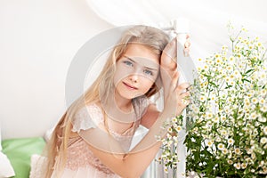 Beautiful little blonde girl with long hair sits on a bed with a bouquet of daisies in a bright white bedroom with a rustic interi photo