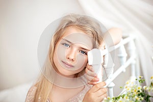 Beautiful little blonde girl with long hair sits on a bed with a bouquet of daisies in a bright white bedroom with a rustic interi