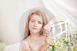 Beautiful little blonde girl with long hair sits on a bed with a bouquet of daisies in a bright white bedroom with a rustic interi