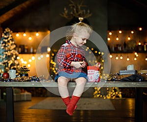 A beautiful little blonde in a traditional plaid shirt opens a chocolate bar from her sweet Christmas gift