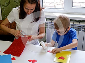 A beautiful little boy and his mother glue a red heart on a yellow leaf. A child makes a greeting card. The life of the home.