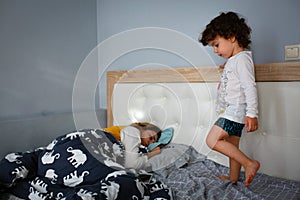 Beautiful little boy playing on the bed in the bedroom next to sleeping mom