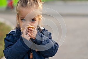 A beautiful little Caucasian girl with blond hair and eating bread eagerly with her hands looks at the camera with sad eyes