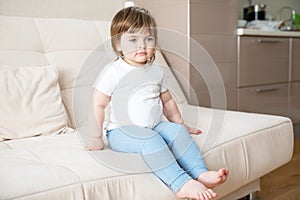 Beautiful little Caucasian girl in white T-shirt and blue pants sitting on light sofa in room.