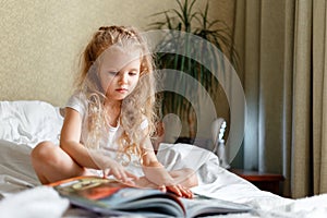 Beautiful little child calm Attentive Child read book on bed. Little blond curly girl Read book in sunny Children& x27;s room