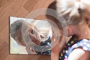 Beautiful little child girl with face painted with color tint in dress looking at her reflection in mirror on floor during self