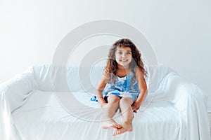 Beautiful little curly girl in a blue dress on the white couch in the room