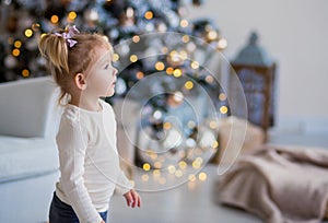 Beautiful little girl with blond hair in a white sweater looks away. Christmas tree in the background, lights, balls