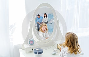 Beautiful little girl with blonde long hair, looking at herself in mirror indoors. Mother and father is in reflection