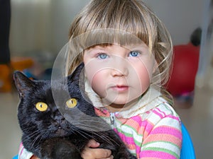 A beautiful little girl with blue eyes is holding a black cat. Friendship with pets.