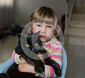 A beautiful little girl with blue eyes is holding a black cat. Friendship with pets.