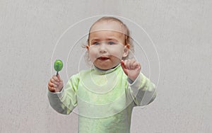 Beautiful little girl with brown eyes and a lollipop in her hands. Happy child in a green sweater eating a green lollipop candy,