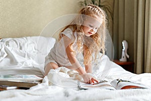 Beautiful little girl Child read book on bed in morning. Little blond curly girl Read book in bedroom sunny Children& x27;s