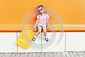 Beautiful little girl child wearing a sunglasses with shopping bags walking in city over colorful orange