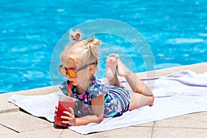 Beautiful little girl, cute toddler with blonde hair in swimwear, sitting at a swimming pool and drinking water melon juice with