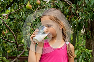 Beautiful little girl drinks milk. Tree with pears in the background