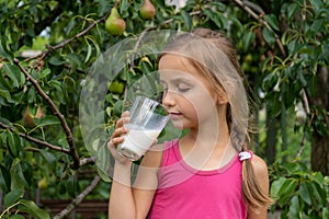 Beautiful little girl drinks milk. Tree with pears in the background