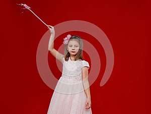 a beautiful little girl in a fairy dress with a magic wand in her hands stands on a red isolated background.