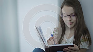 Beautiful little girl with glasses writes in his diary at window.