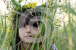 Beautiful little girl with green eyes and a colorful garlang made of wild flowers on her head sits in high green grass on a meadow