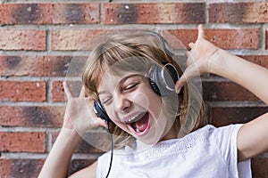 beautiful little girl with headphones at home listening to music and singing, technology and music concept. Brick background.