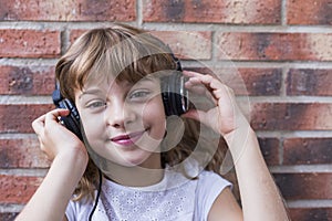 Beautiful little girl with headphones at home listening to music and smiling, technology and music concept. Brick background