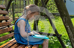 Beautiful little girl is learning to draw and write while sitting on a swing on a sunny day. Side view