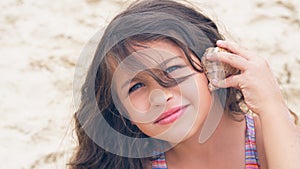 Beautiful little girl with long curly hair listening to sea music in cockleshell on the beach.