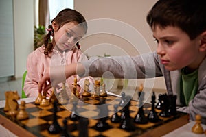 Beautiful little girl looking at her brother moving his chess piece over chessboard, making his move while playing chess game