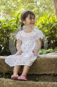 Beautiful little girl looking at something while sitting on the concrete edge of a public garden